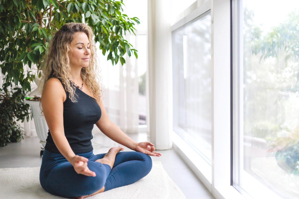 Woman yoga meditation sitting in lotus position near panorama window with houseplant at home